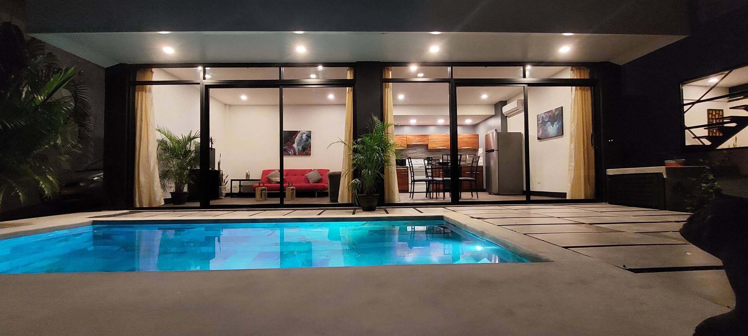 A swimming pool with a red sofa and dining area in the background.