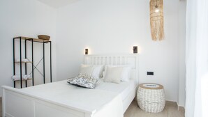 Quarto branco com cama, mesas de cabeceira, luminária pendente e uma pequena cesta redonda de vime.