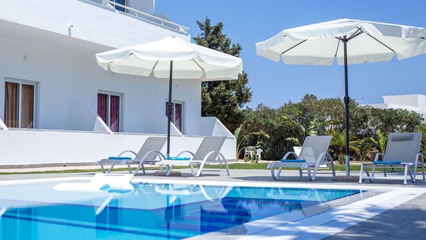 A swimming pool with lounge chairs and umbrellas, a white building with windows, and greenery in the background. - 14 Ground Floor (Faliraki)