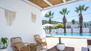 A covered outdoor seating area with wicker furniture, a potted plant, and a view of a pool and buildings in the background. - 10 Ground Floor (Faliraki)