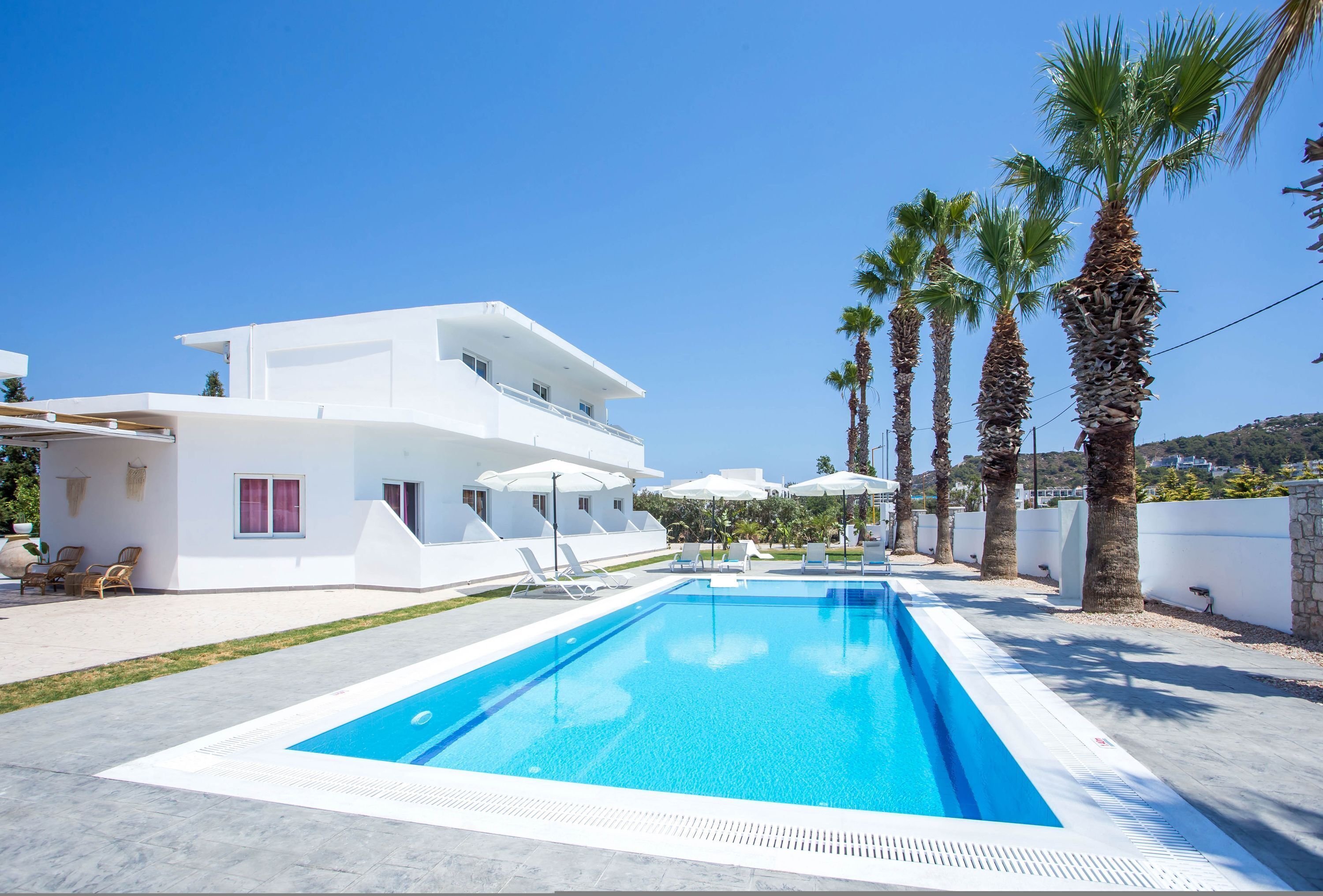 A modern white building with a swimming pool, palm trees, and a clear blue sky.