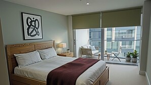 Une chambre avec un grand lit, une table de chevet, une lampe, une chaise, une petite table et un balcon agrémenté de plantes en pot.
