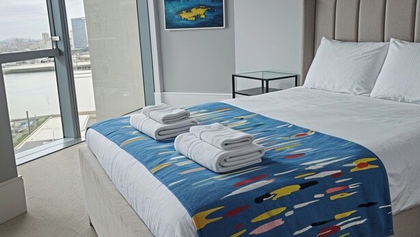 Une chambre d’hôtel moderne avec un grand lit, une vue sur la ville, un tableau au mur et une porte vitrée donnant sur un balcon.