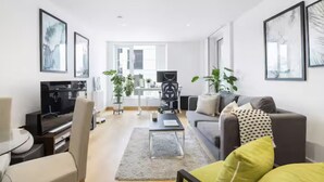 Ein modernes Wohnzimmer mit einer Couch, Couchtisch und Fernseher. An den Wänden hängen gerahmte Bilder, und ein Schreibtisch wird von einem Stuhl ergänzt.