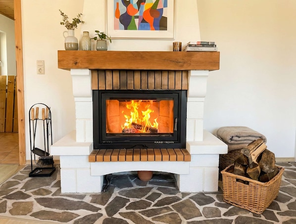 Une cheminée avec un manteau de couleur blanche, un tableau abstrait encadré au-dessus, et un panier de bois de chauffage sur la droite.