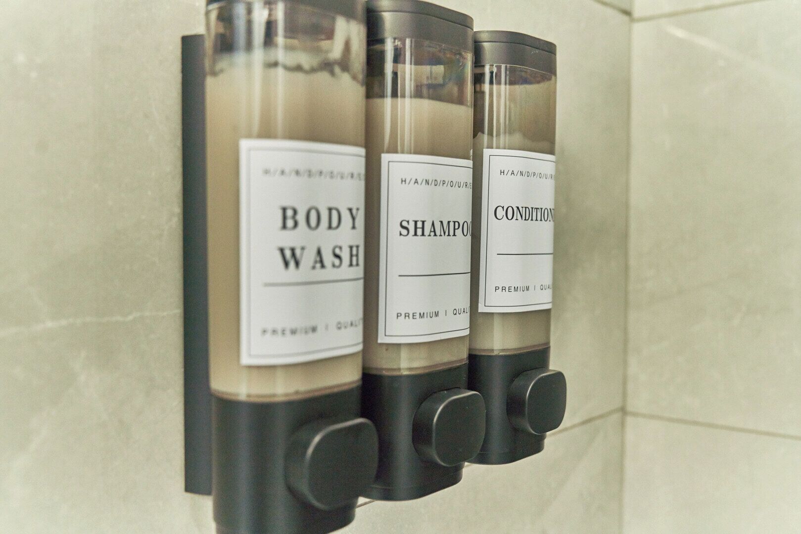 Trois flacons de gel douche, de shampoing et d’après-shampoing, placés sur un distributeur mural.
