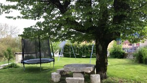 A trampoline under a tree, a stone bench, and a swing set in a grassy yard.