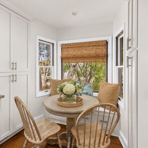 A corner dining area with a round table, two wooden chairs, a window with a bamboo blind, and a view of trees. - 806 Carolina - 1 Block to Beach/Ocean! (Isle of Palms)