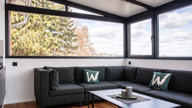 A modern living room with a dark grey sectional sofa, a small coffee table, and large windows offering a view of trees and the sky. - Vacation home in secluded location near Eifel National Park (Mechernich)