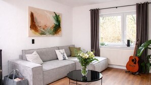 A living room with a grey sofa, a glass coffee table, a guitar, and a painting on the wall. - Vacation home in secluded location near Eifel National Park (Mechernich)