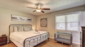 Une chambre à coucher comprenant un lit, un ventilateur de plafond, une fenêtre avec des stores, une table de chevet et une œuvre d’art accrochée au mur.