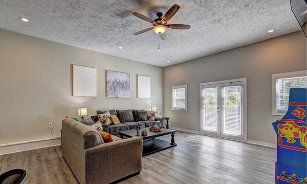 A spacious living room with a ceiling fan, a sofa, a coffee table, and a large window with blinds. - Chop House - King Bed, Smart TVs & Backyard Fun! (Douglasville)