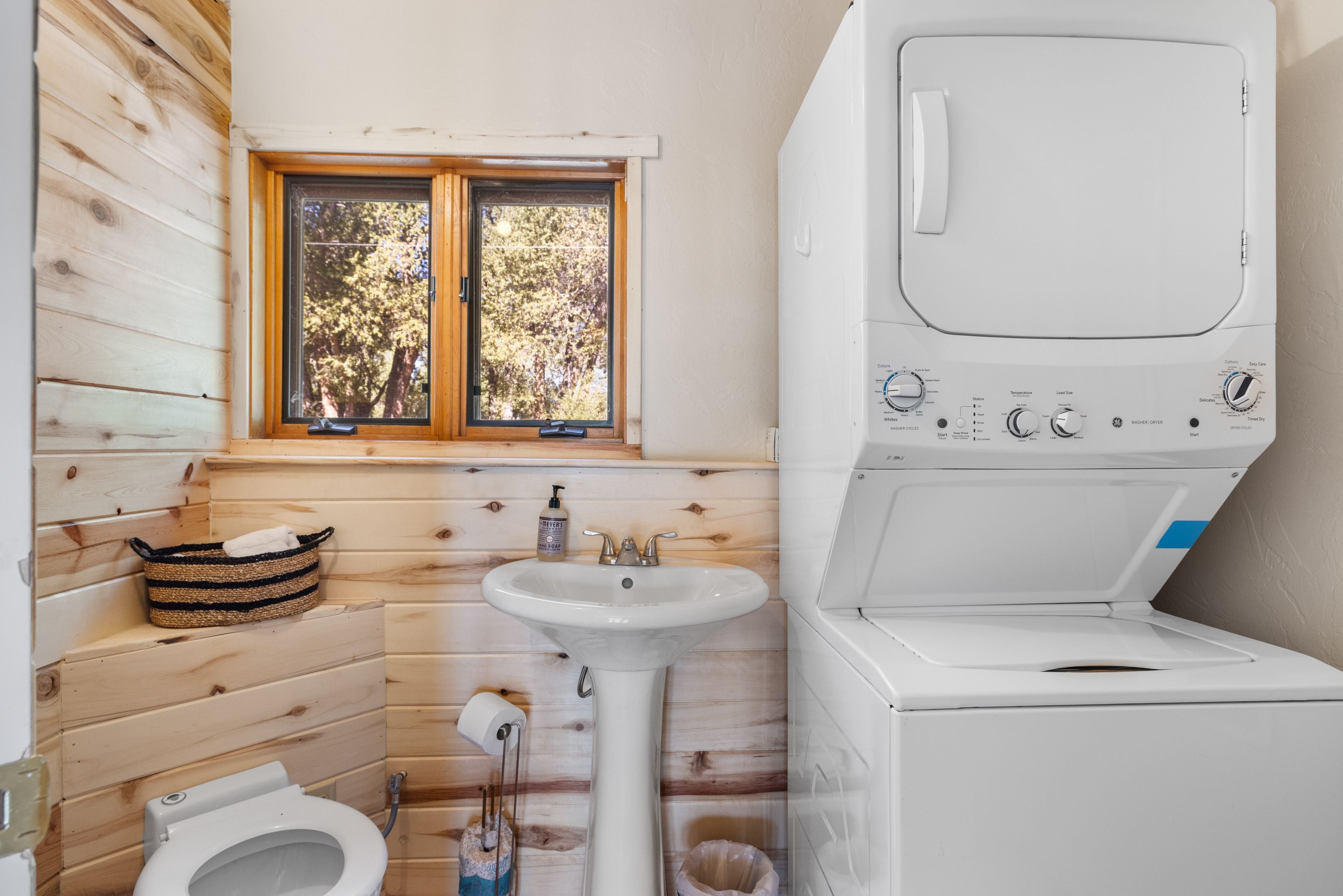 A compact bathroom with a wooden interior, a window, a sink, a toilet, and a washing machine.