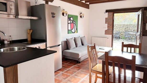 A compact kitchen with a refrigerator, microwave, and sink. There's a dining area with a table and chairs, and a sofa. The room has exposed wooden beams and a view through a window.