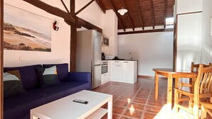 A living area with a sofa, a coffee table, a dining table with chairs, a kitchen in the background, and a ceiling with wooden beams.