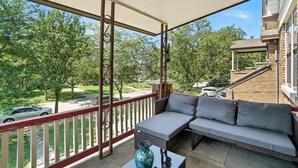 Een overdekt balkon met een bank en een tafel, met uitzicht op een woonstraat en groen.