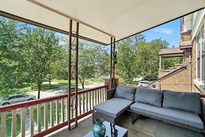 A covered balcony with a sofa and a table, overlooking a residential street and greenery. - 3BR, Quiet Street, 10 Min DTW (Cincinnati)
