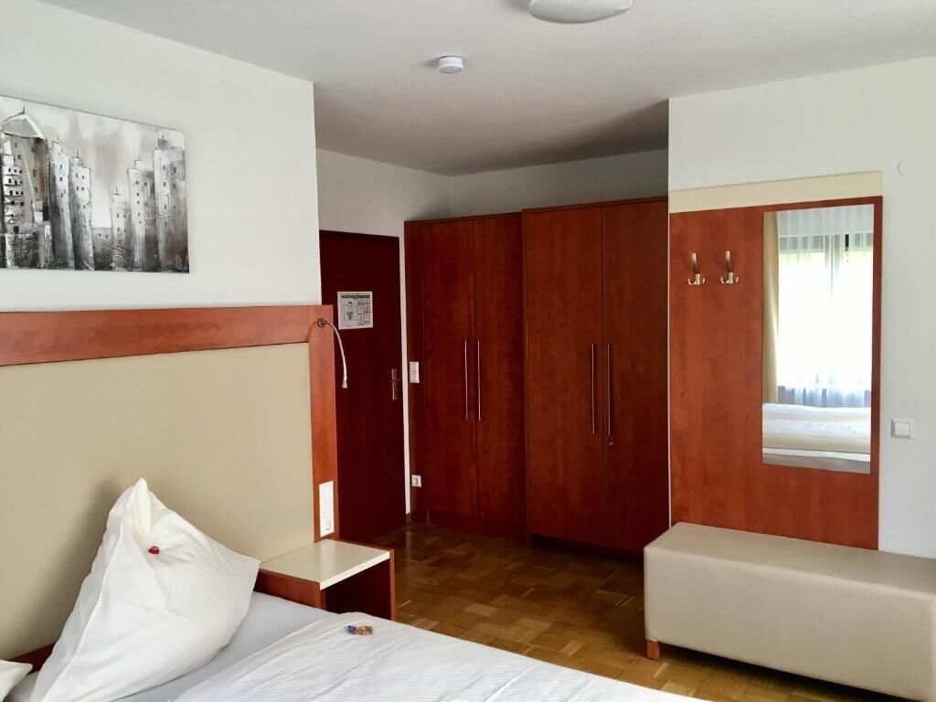 A hotel room with a bed, a small bedside table, a wooden wardrobe, a window with a view, and a painting on the wall.