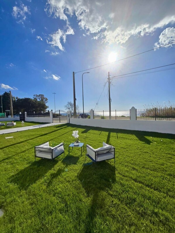 A grassy outdoor area with three white lounge chairs and a small table, surrounded by a low fence and street lamps.