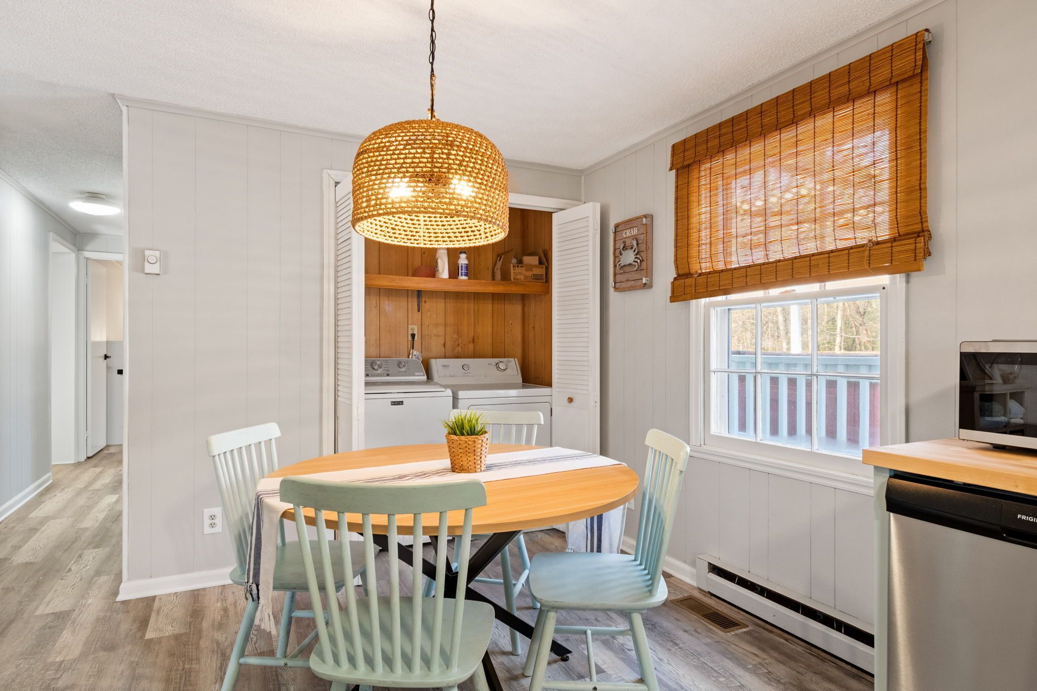 A dining area with a wooden table, two chairs, a hanging light fixture, and a kitchen in the background.