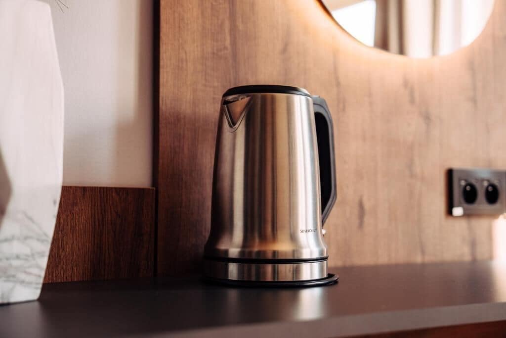 A stainless steel kettle on a dark countertop with a wooden panel and a circular mirror in the background.