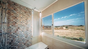Un baño con pared de mosaicos, ducha, banco y ventana grande con vista a un paisaje rural.