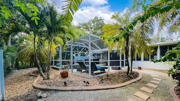 Un espace extérieur abrité par des parois de verre, comprenant des sièges et une piscine, le tout entouré de palmiers et bordé d’un chemin de gravier.
