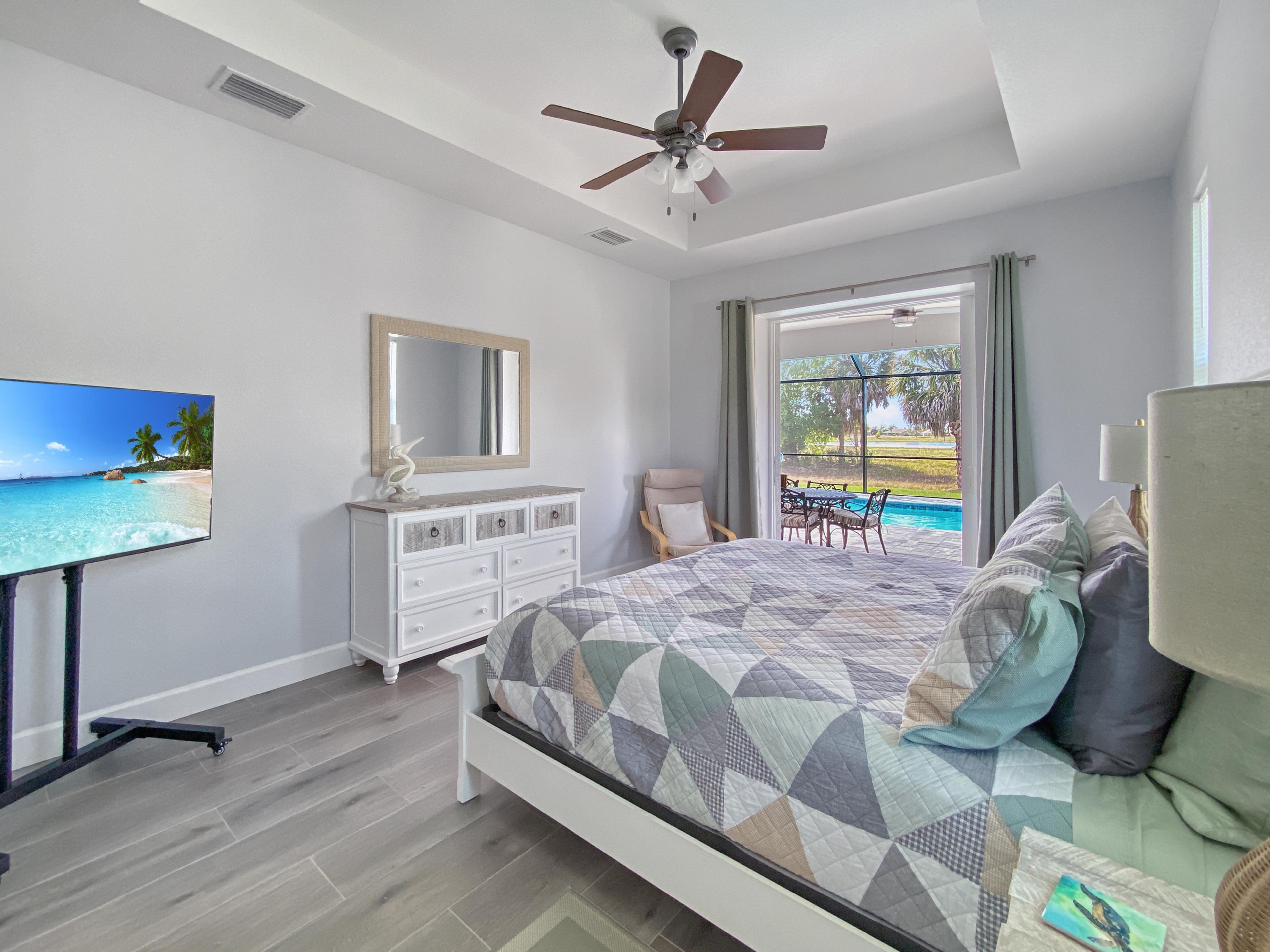 Une chambre avec un lit, une commode, une télévision, un ventilateur de plafond et une vue sur une piscine.