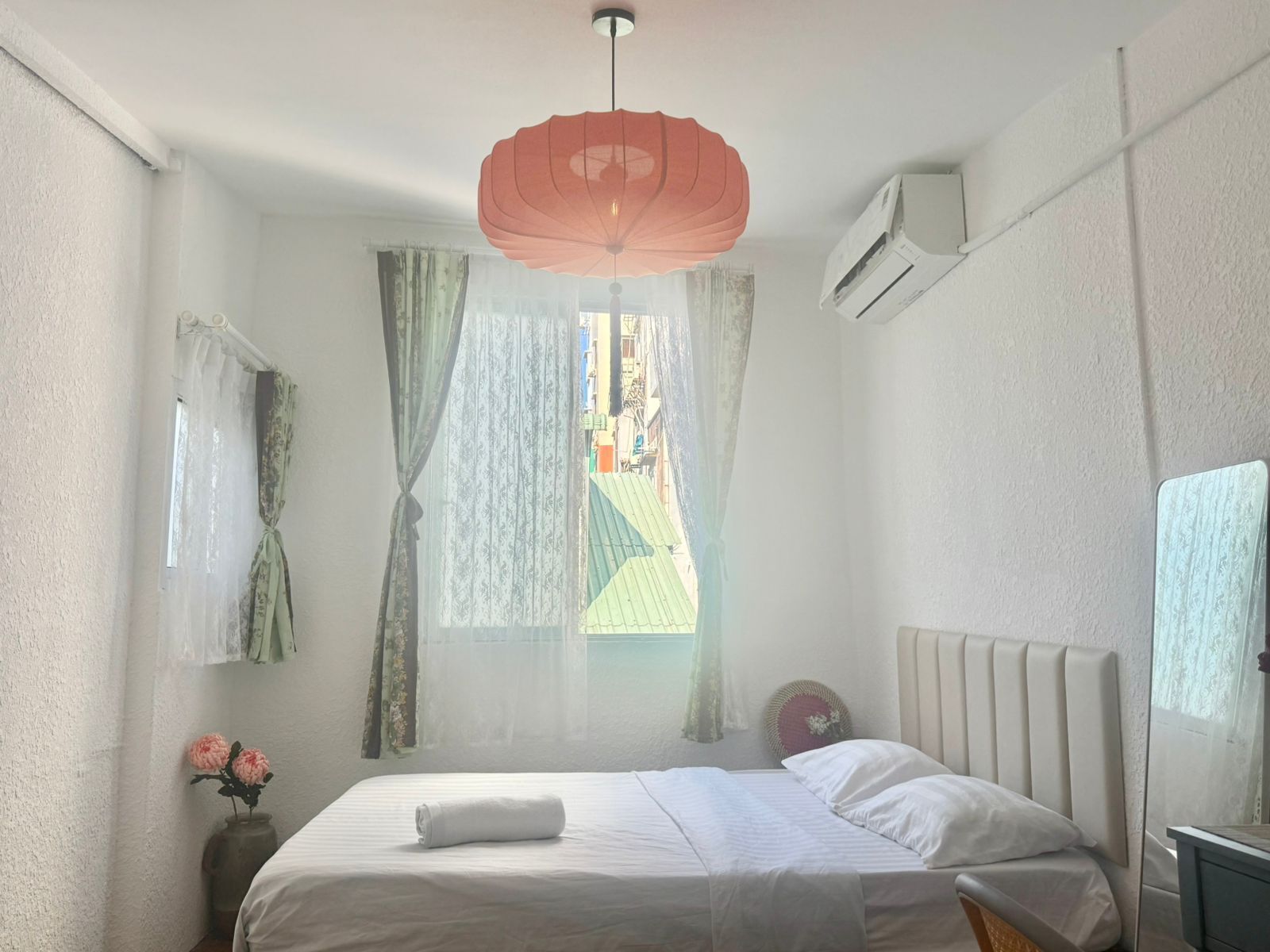 A bedroom with a bed, a hanging light fixture, a window with curtains, and an air conditioning unit.
