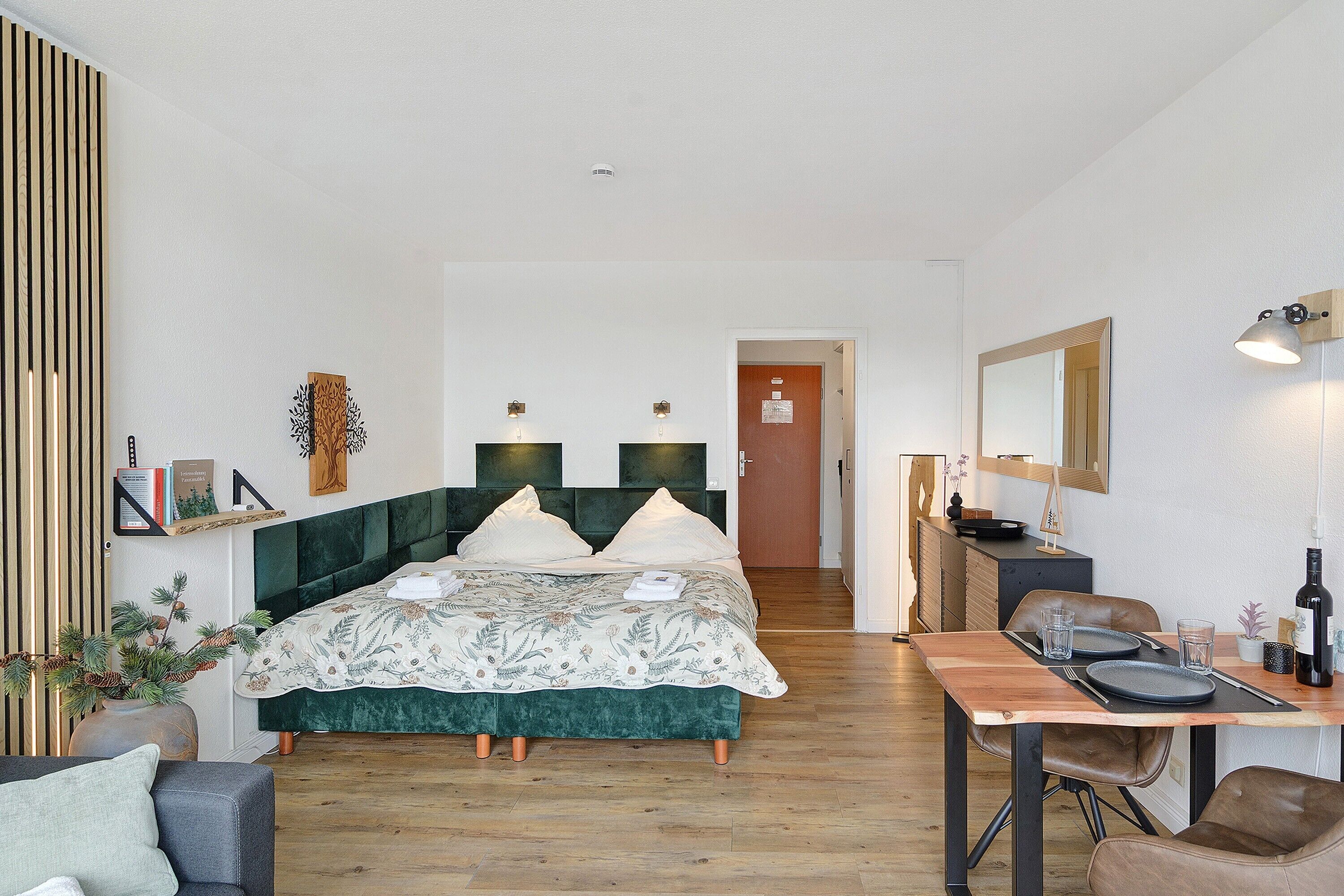 A modern bedroom with a bed, a dining table with chairs, a mirror, and a small plant.