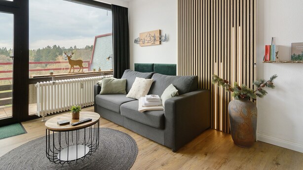 A modern living room with a grey sofa, a round coffee table, a large window with a view of a deer, a radiator, and a tall vase with greenery. - Apartment 'Panoramablick Hahnenk' with Sea View, Wi-Fi and Air Conditioning (Goslar)