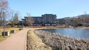 A modern building with a flat roof, a paved walkway beside a body of water, and a grassy area with trees. - Apartment 'Panoramablick Hahnenk' with Sea View, Wi-Fi and Air Conditioning (Goslar)