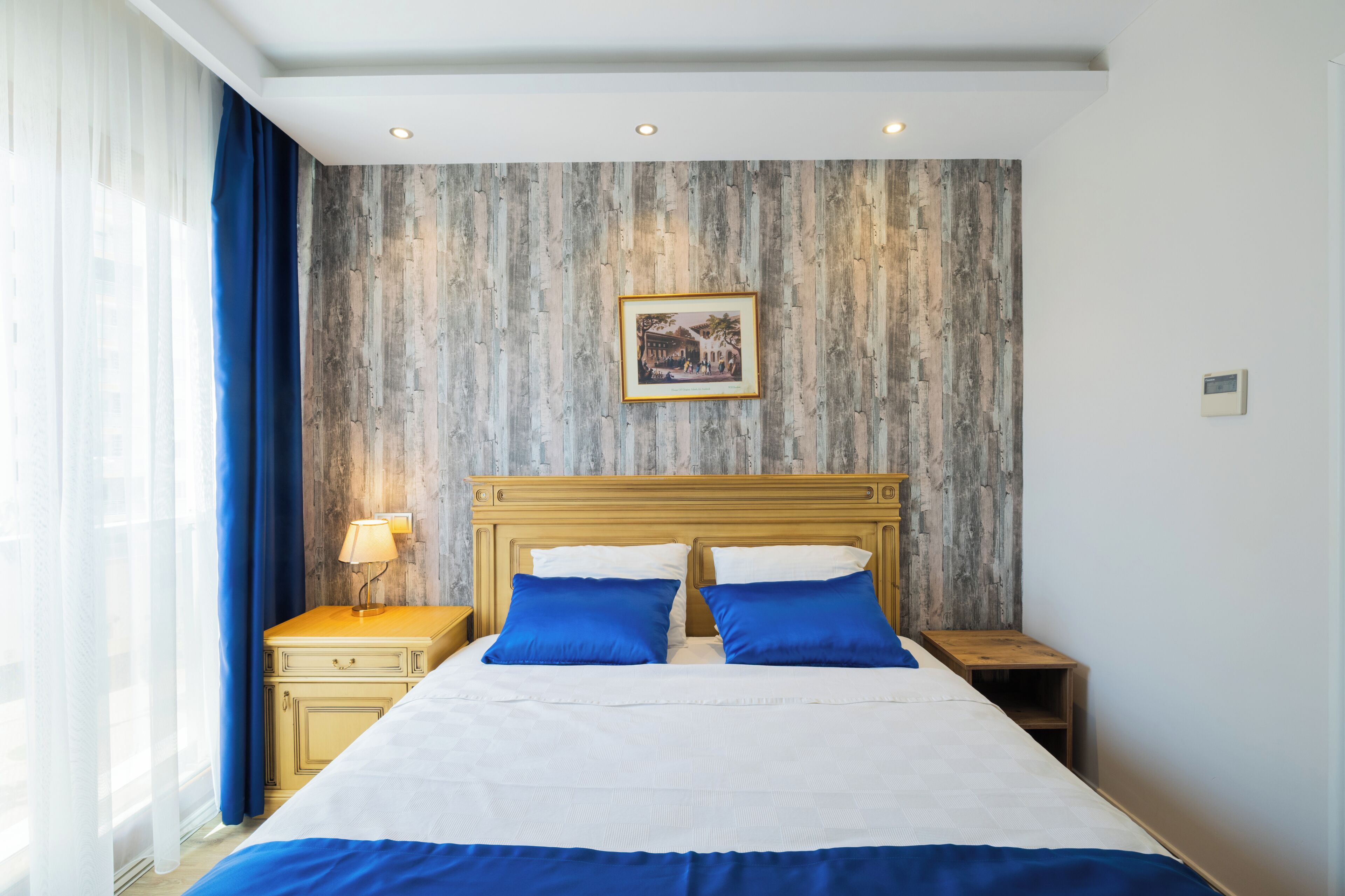 Habitación de hotel con cama, dos mesitas de noche, un velador, un cuadro colgado en la pared y una ventana con cortinas.