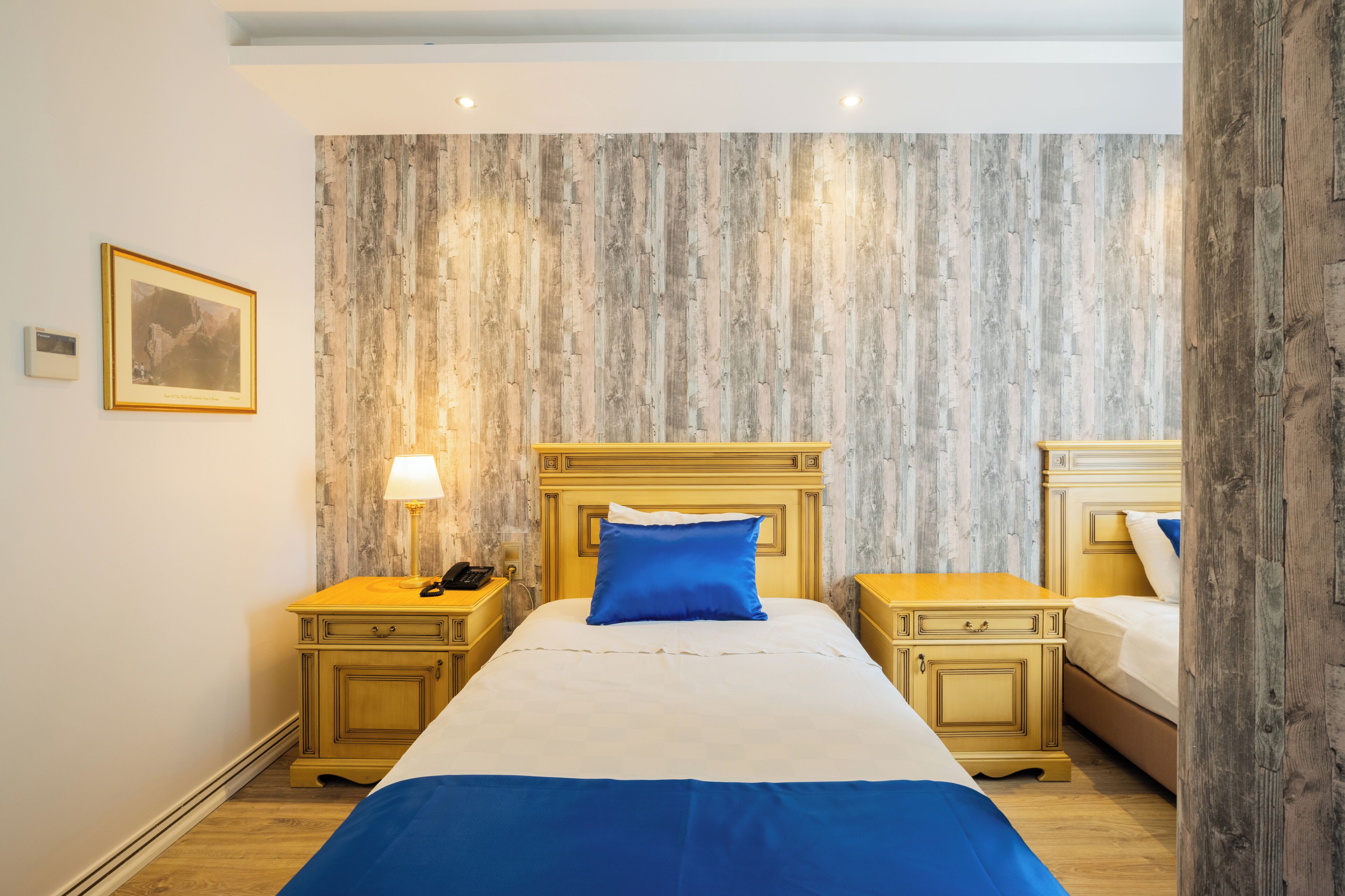 Hotellihuone, jossa on sänky, kaksi yöpöytää, yölamppu ja seinällä oleva kehystetty kuva.