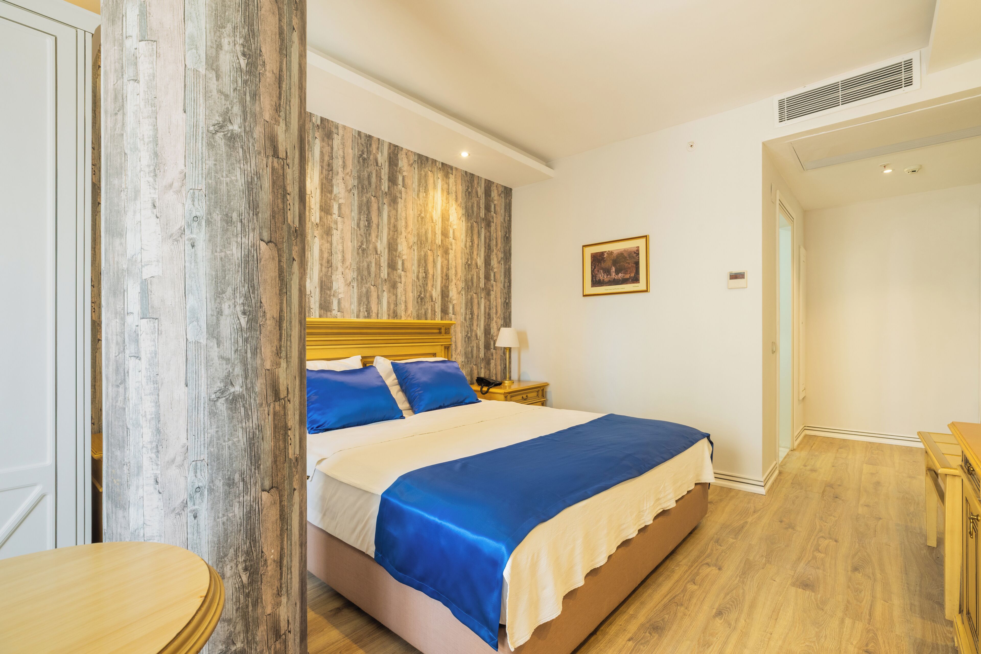A bedroom with a wooden headboard, a bed with blue bedding, a bedside table with a lamp, and a small wooden table.