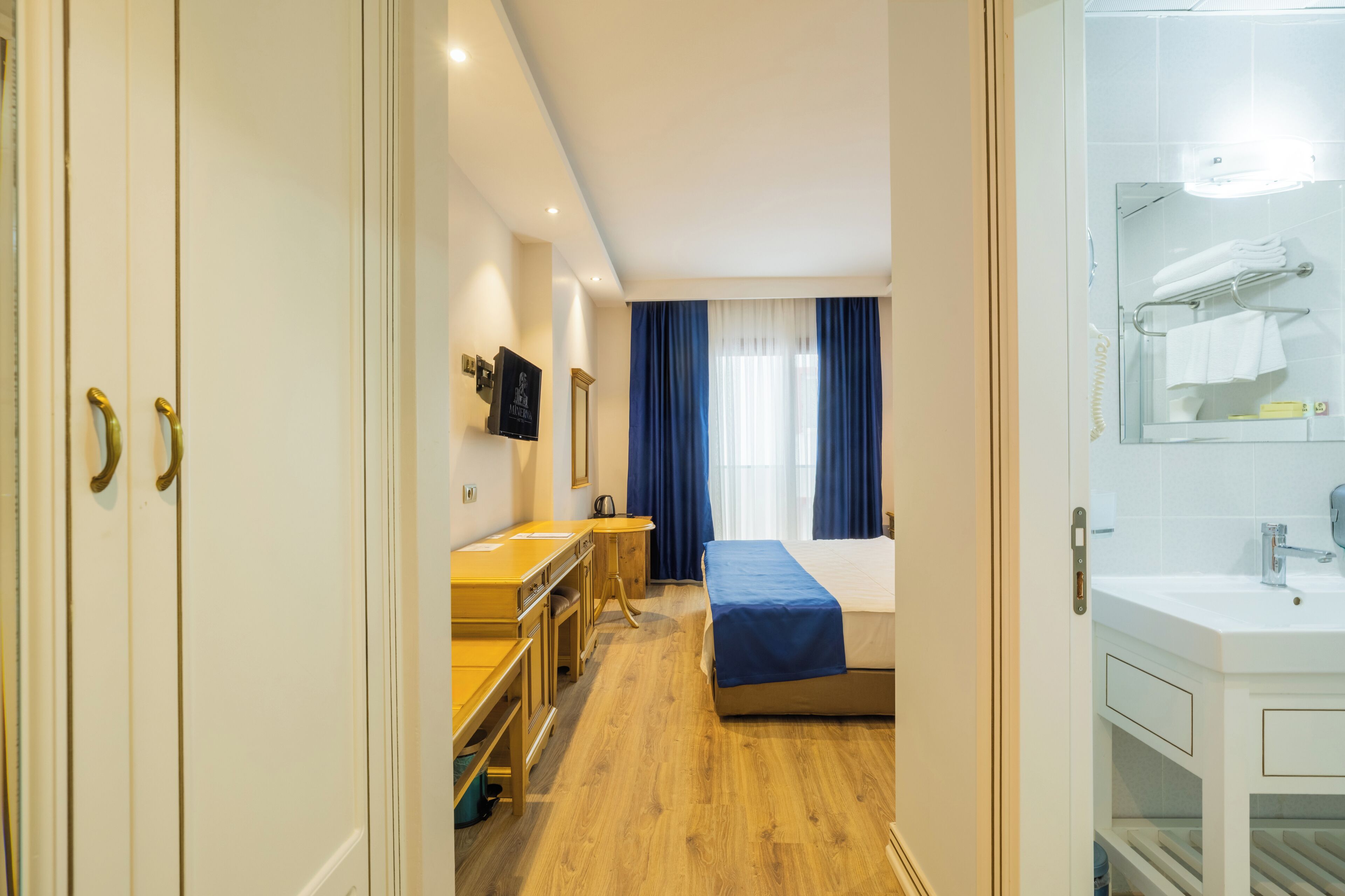 Hotellihuone, jossa on sänky, työpöytä, peili sekä kylpyhuone, jossa on pesuallas ja pyyheteline.