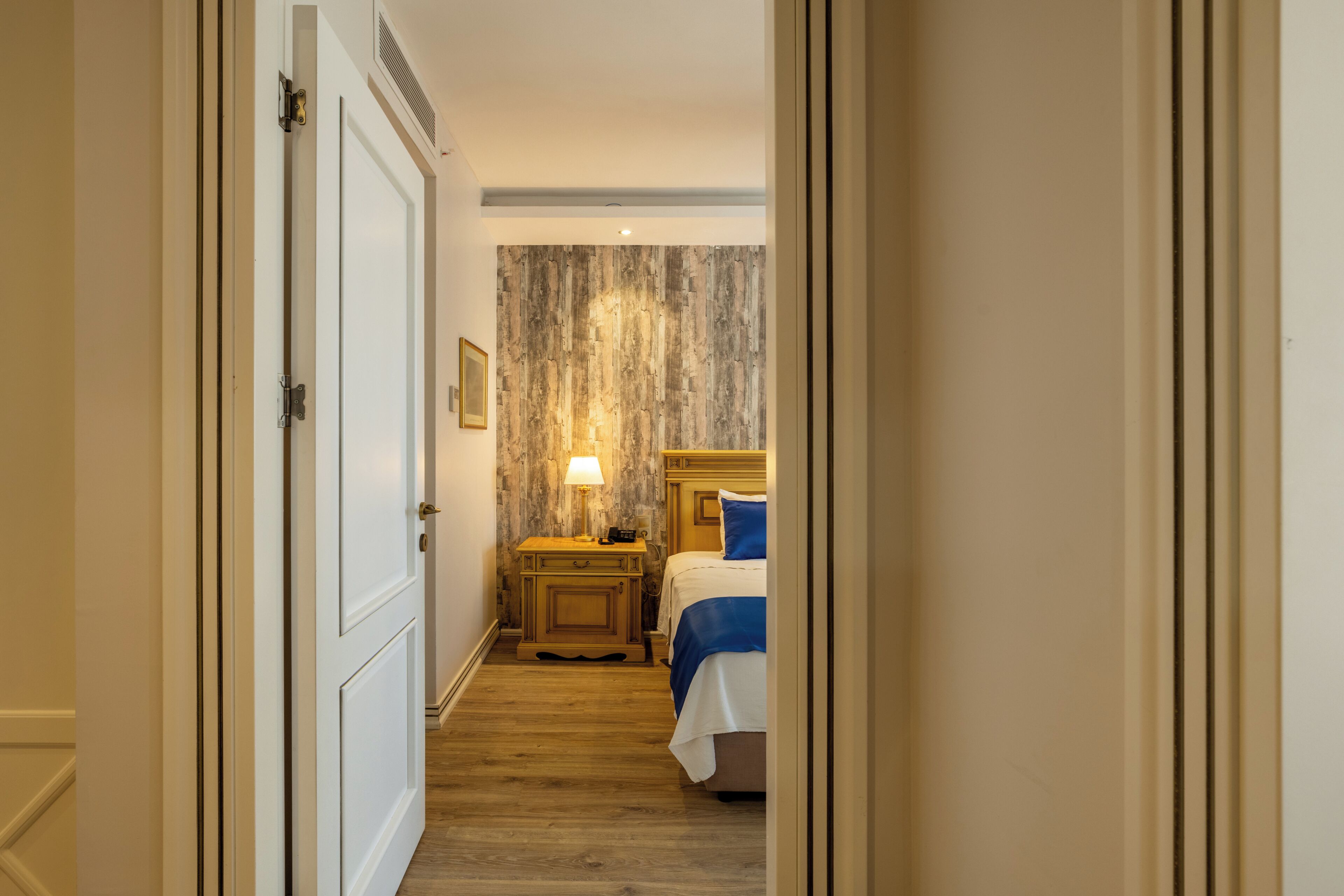 A hotel room with a wooden floor, a bed with blue bedding, a nightstand with a lamp, and a textured wall.