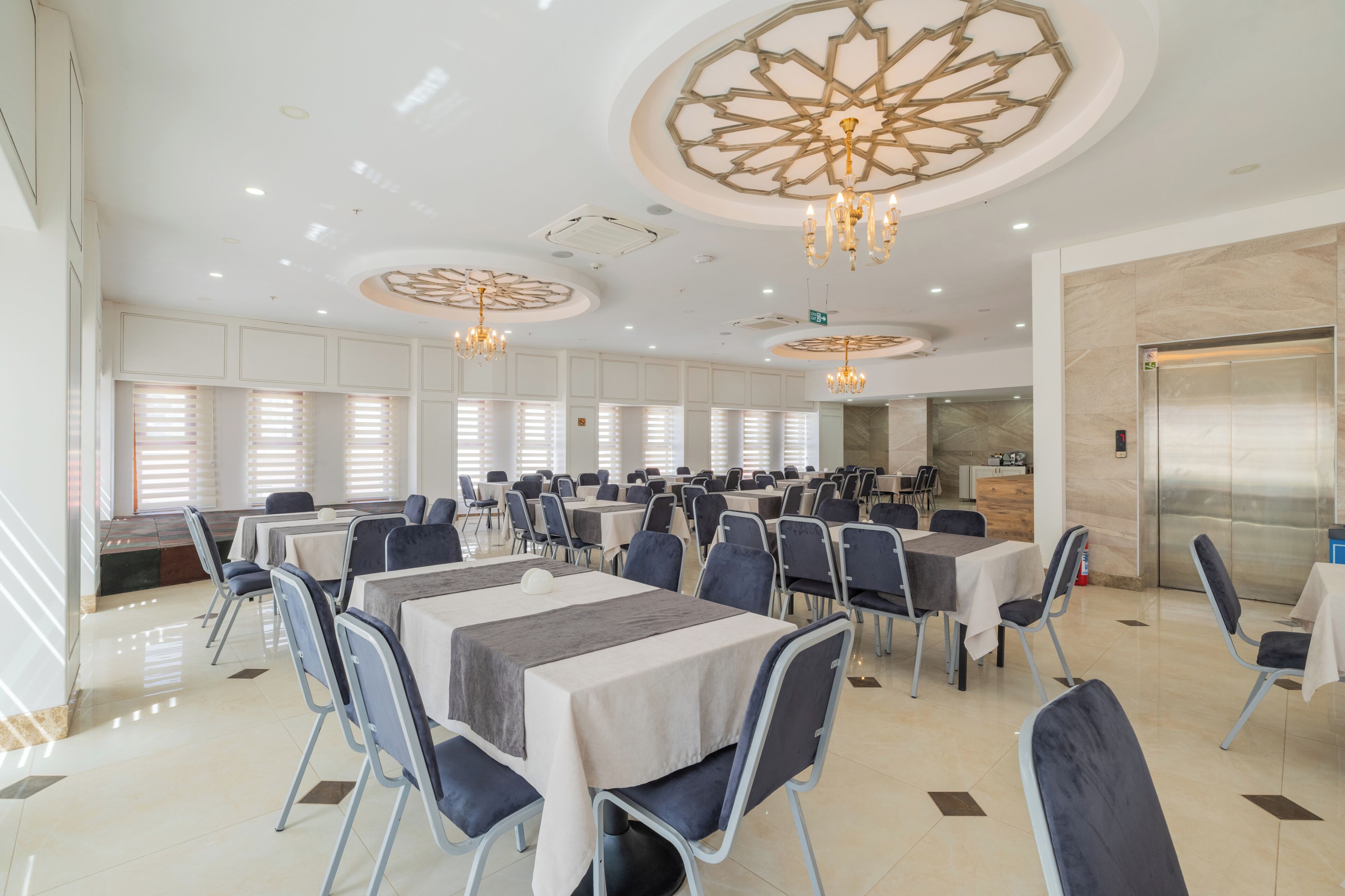 A spacious dining area with multiple tables and chairs, a central chandelier, and a modern ceiling design.