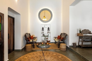 A lobby with a round wooden table, two armchairs, a mirror, and a wall-mounted shelf with decorative items. - Staymaster Villa Anasoh 4BHK (Vagator)