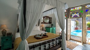 A four-poster bed with a canopy, a bedside table with a lamp, a mirror, and a view of an outdoor swimming pool. - Staymaster Villa Anasoh 4BHK (Vagator)
