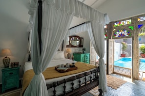 A four-poster bed with a canopy, a bedside table with a lamp, a mirror, and a view of an outdoor swimming pool. - Staymaster Villa Anasoh 4BHK (Vagator)