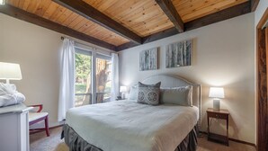 Een slaapkamer met een bed, twee bedlampen, een raam met gordijnen en uitzicht op bomen.