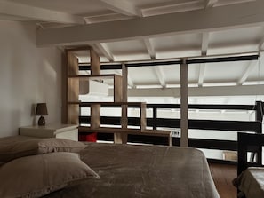 Ein Schlafzimmer mit Bett, Nachttisch, Lampe, einer Holzregaleinheit und Blick durch große Fenster.
