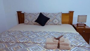 Una cama con una colcha estampada, un almohadón negro y una mesita de noche de madera con una lámpara.