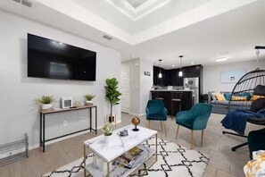 A modern living room with a flat-screen TV, a dining table, teal chairs, and a kitchen in the background. - Furnished 2BR/2BA Monthly • Comfy King • Workspace + Laundry • Near Hospitals (Edinburg)