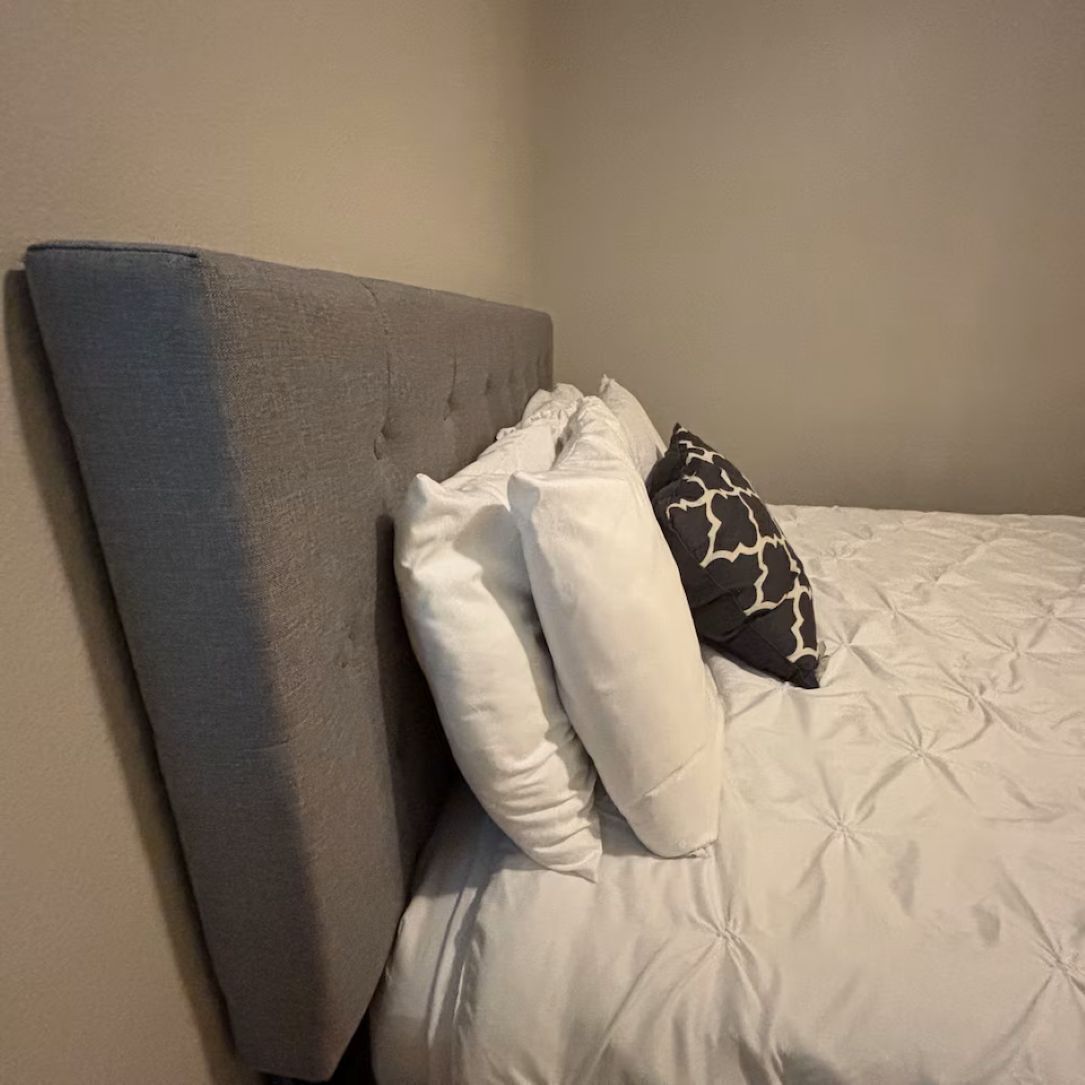 Cama com cabeceira cinza, almofadas brancas e estampadas, e edredom branco.