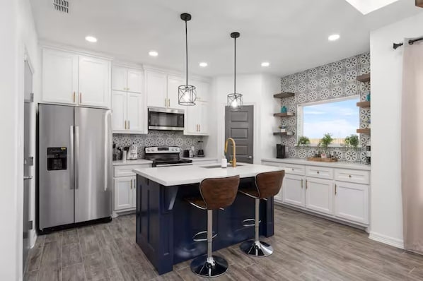 A modern kitchen with white cabinets, stainless steel appliances, a central island with bar stools, pendant lighting, and a patterned backsplash. - The Spacious Nest 3BR/2BA,King Bed,Near Hospital (Edinburg)