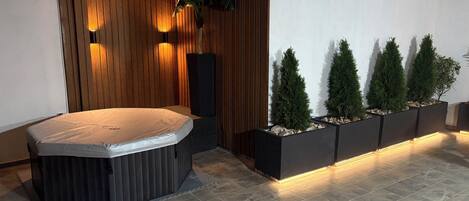 A modern spa area with a hot tub, wooden panel walls, and a row of planters with coniferous trees.