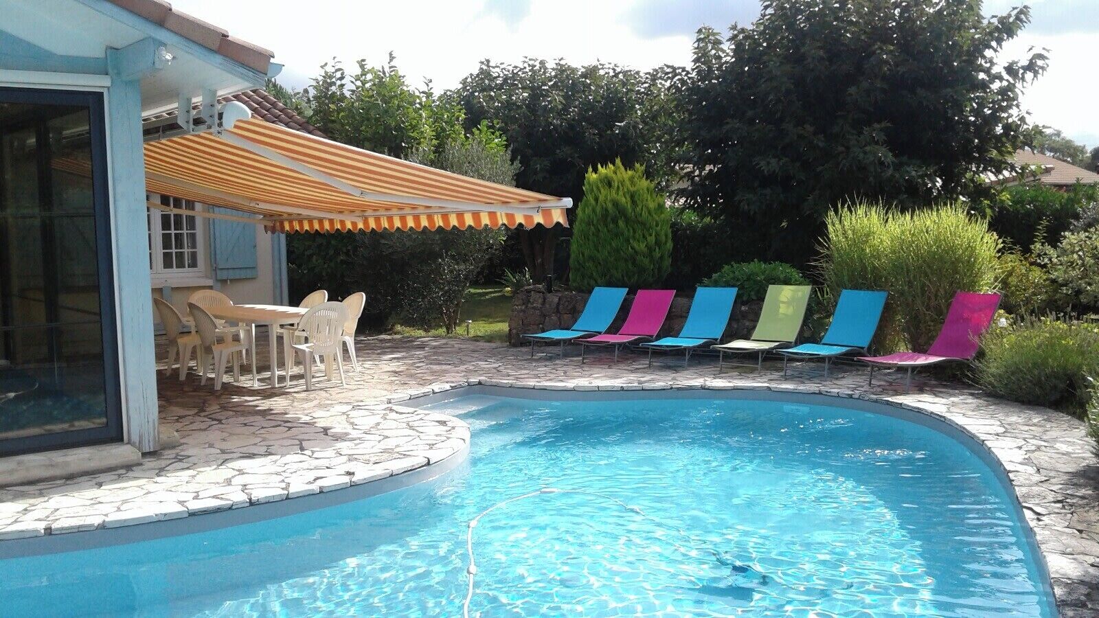 A swimming pool with a tiled deck, a covered patio with chairs, and colorful lounge chairs.