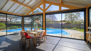 Eine überdachte Terrasse mit Esstisch und Stühlen, großen Fenstern und Blick auf einen Pool.